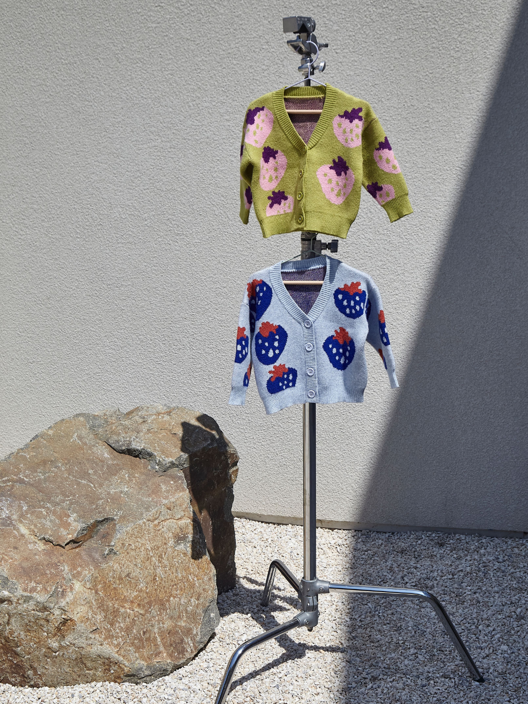 Two kids v-neck cardigans, shown on hangers, hanging from a metal photography stand, in an outdoor space with a gravel floor, a large rock and grey wall. The lower cardigan is light blue with an all-over pattern of large blue strawberries with a red leaf. The cardigan hanging directly above is green with a pink strawberry with a purple leaf.