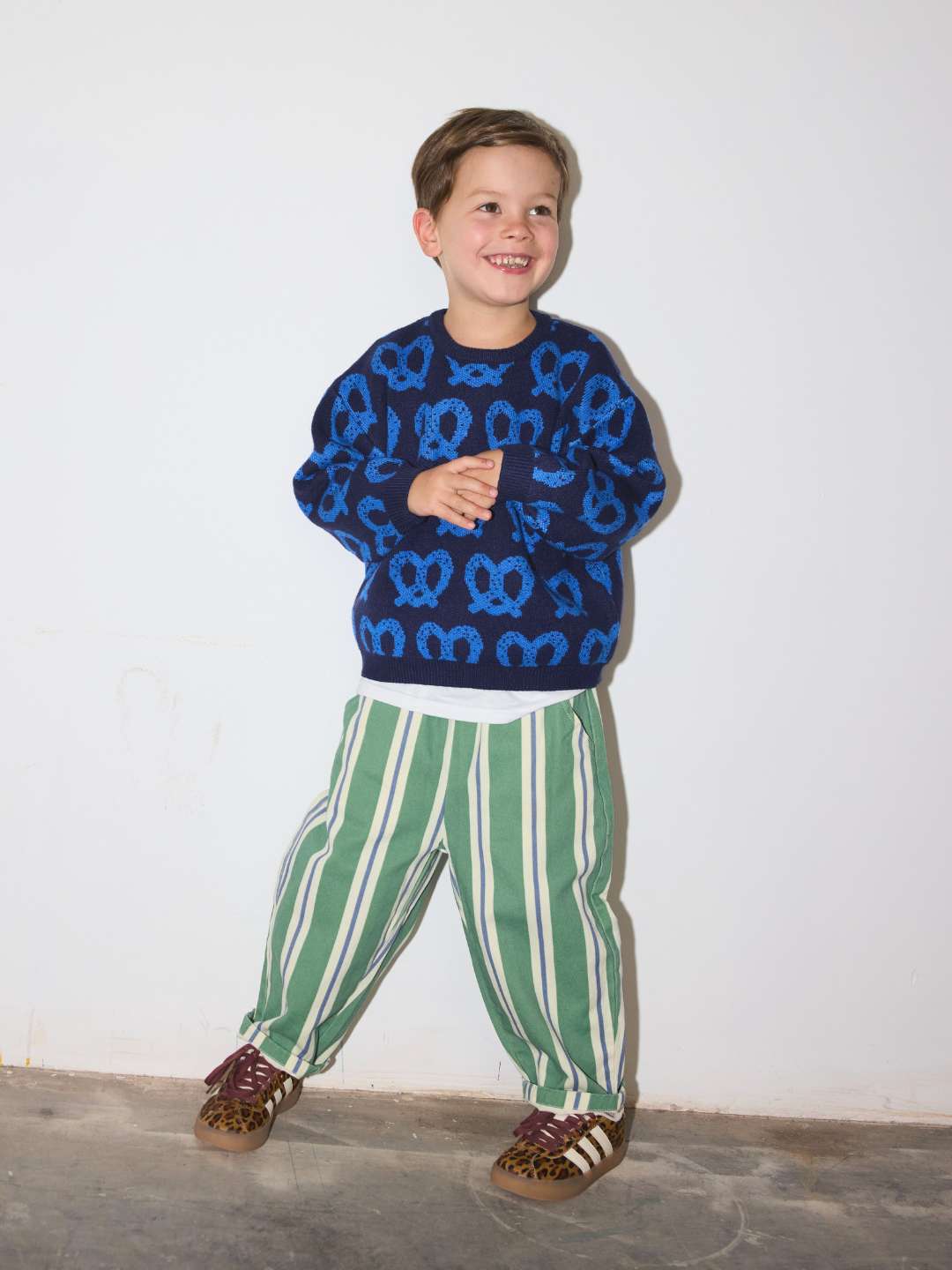 Blue | A young boy wearing a blue and navy pretzel patterned crewneck sweatshirt, green and white vertically striped pants, and brown sneakers, standing against a plain white wall.