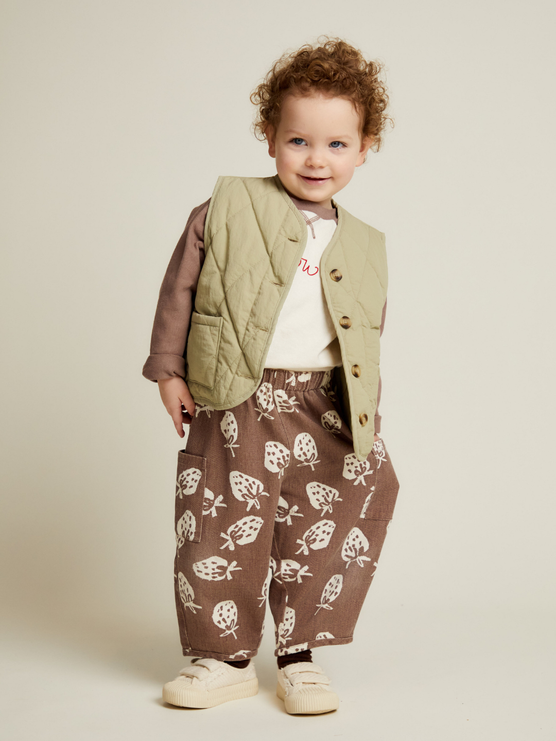 A young child with curly hair stands smiling against a plain background, wearing the EMERSON CHOC STRAB TROUSER paired with a light green vest, long-sleeve shirt, and beige shoes.