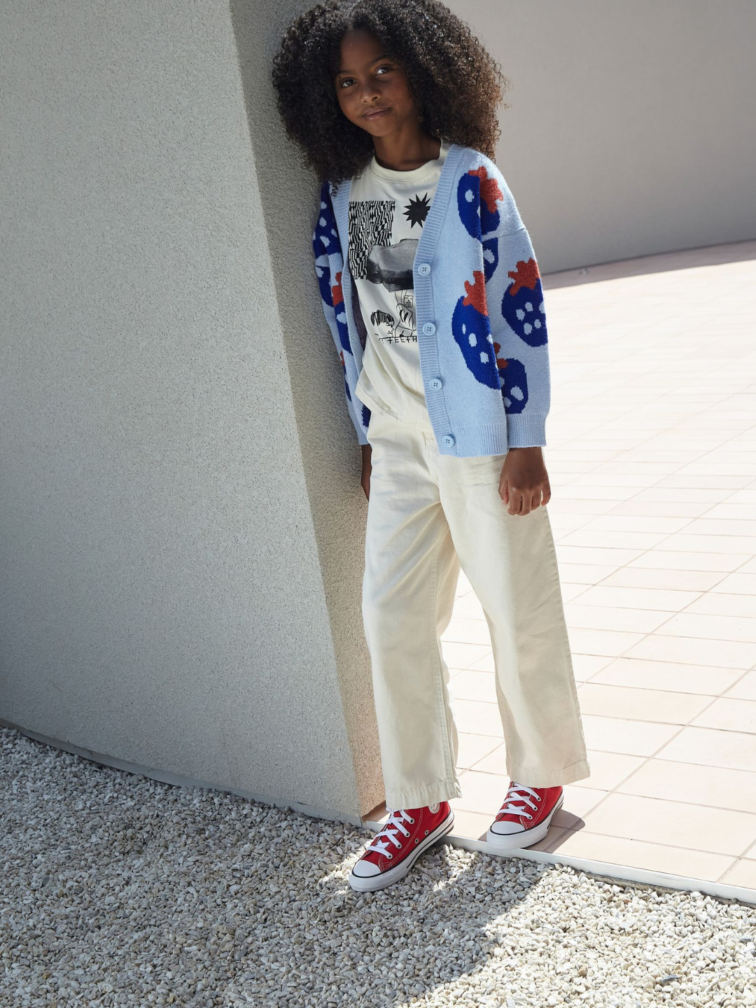 A girl wearing a kids v-neck cardigan in light blue with an all-over pattern of large blue strawberries with a red leaf. She is leaning against a concrete wall in a paved outdoor space with gravel, and wearing a white t-shirt with a black graphic, white jeans, and red hi-top sneakers.