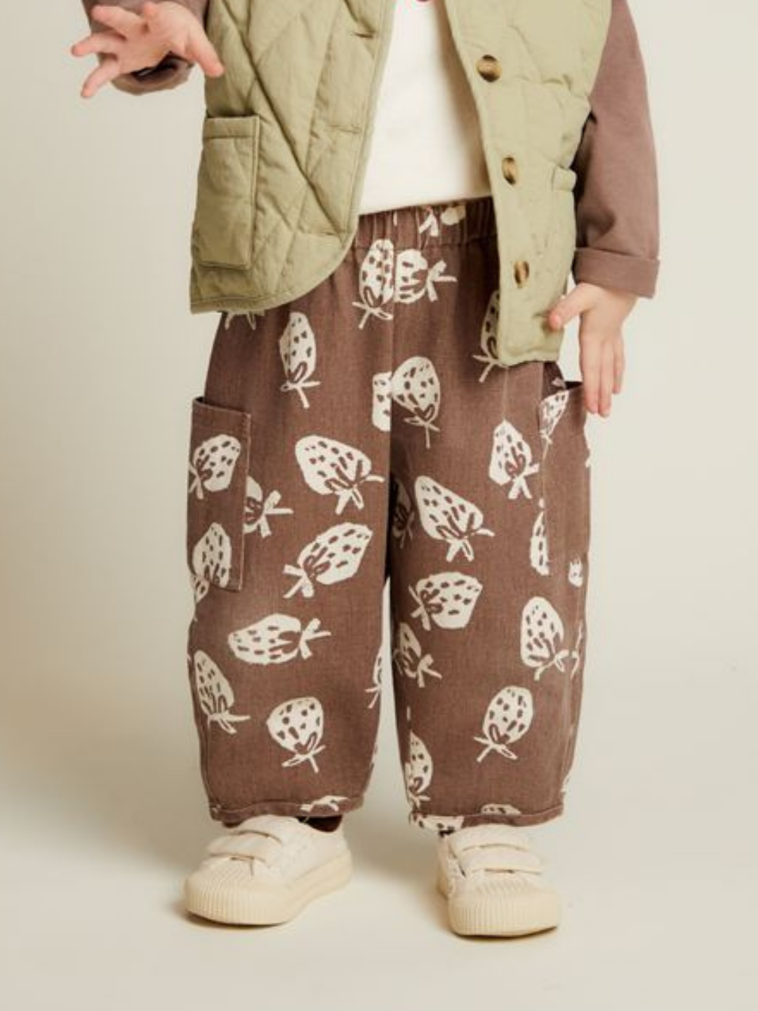 A child wearing the EMERSON CHOC STRAB TROUSER, a cream shirt, light green vest, and beige sneakers stands with one hand raised.
