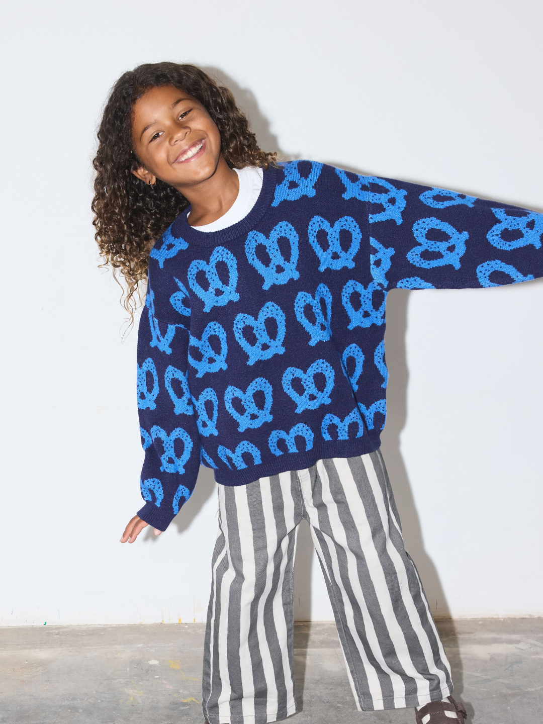 Blue | A smiling girl with curly hair wearing a navy blue crewneck sweater with a bright blue pretzel pattern, paired with gray and white striped pants, posing against a plain white wall.