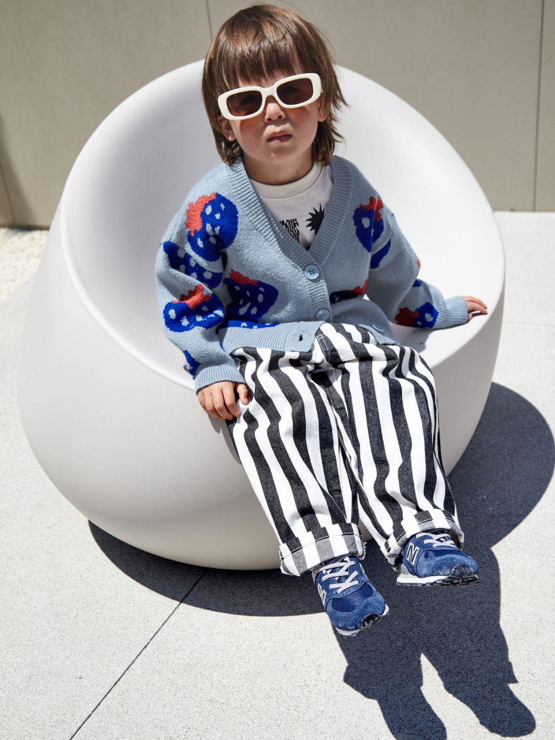 A child wearing a kids v-neck cardigan in light blue with an all-over pattern of large blue strawberries with a red leaf. He is seated on a round white chair in a paved outdoor space.
