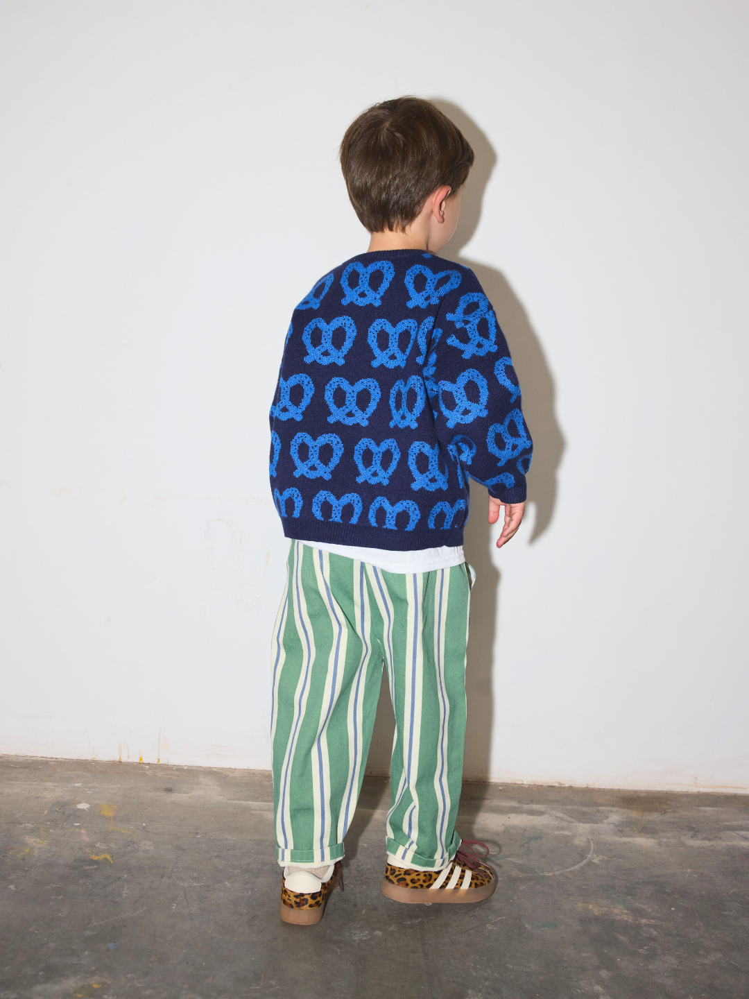 Blue | A child standing with their back to the camera, wearing a navy blue sweater with a blue pretzel pattern, green and white striped pants, and leopard print sneakers, set against a plain white wall and concrete floor.