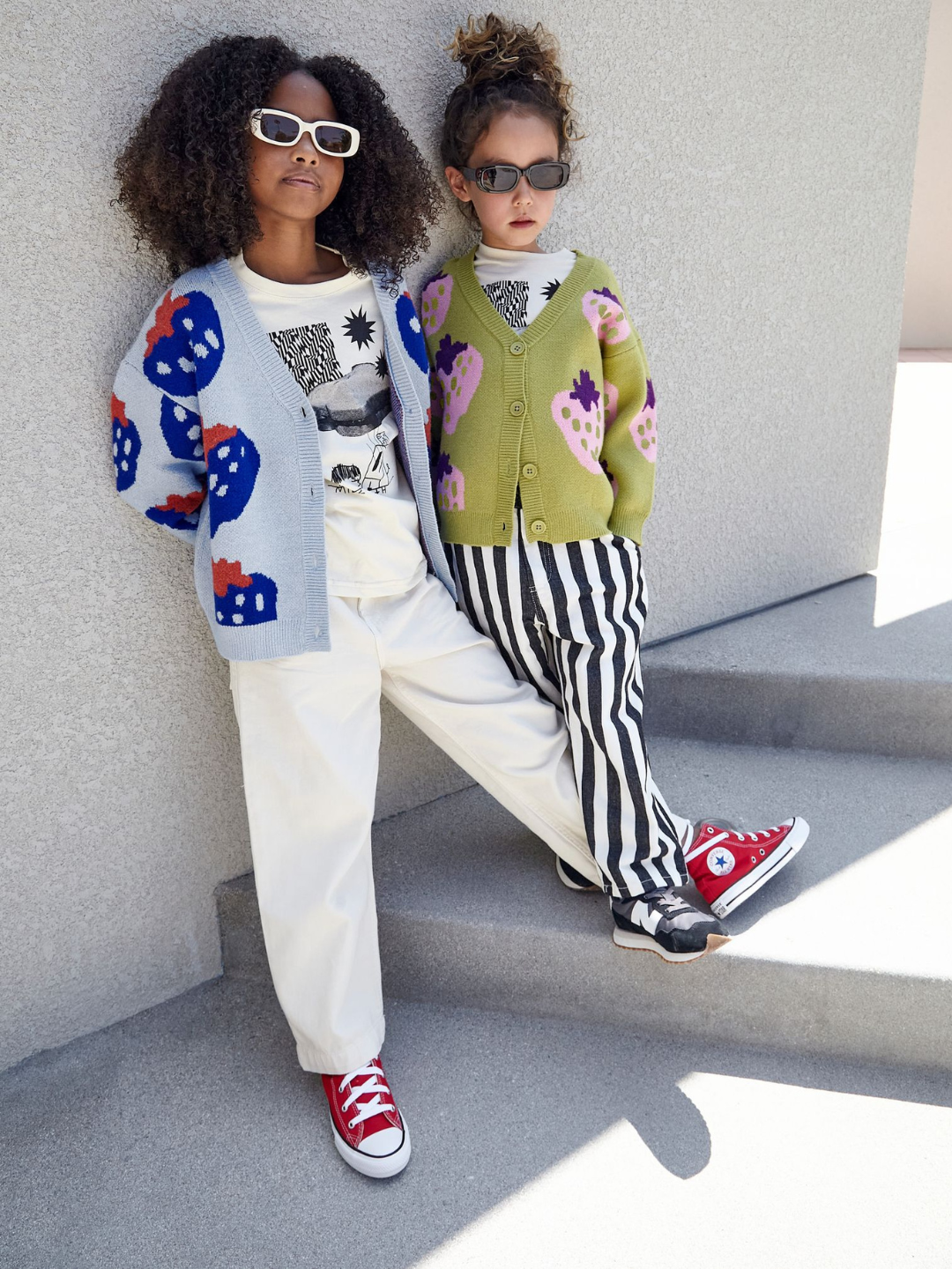 Two kids standing against a grey wall. One girl wears a blue cardigan patterned with darker blue strawberries, worn with a white graphic tee and white jeans, white rectangular sunglasses, and red hi-top sneakers. The other girl wears the same cardigan style, but in green with pink strawberries. She wears it with black rectangular sunglasses, a white graphic tee, black and white striped jeans, and grey sneakers.