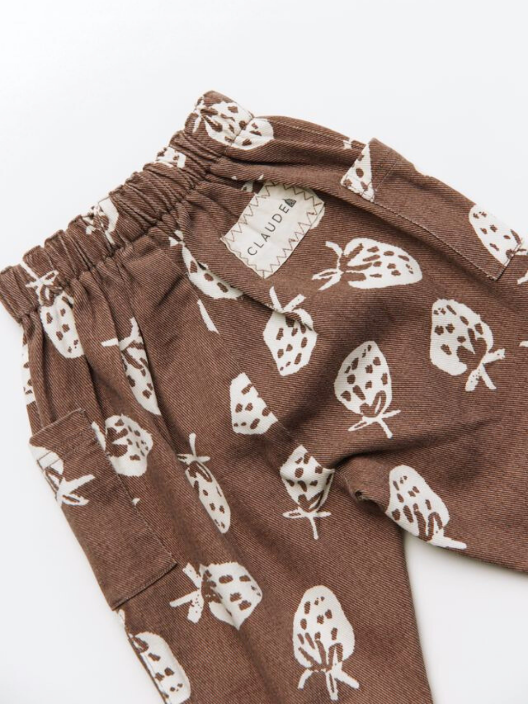 EMERSON CHOC STRAB TROUSER: Brown fabric pants featuring a white strawberry print, elastic waistband, side pocket, and a sewn-in "CLAUDE" label with zigzag stitching.