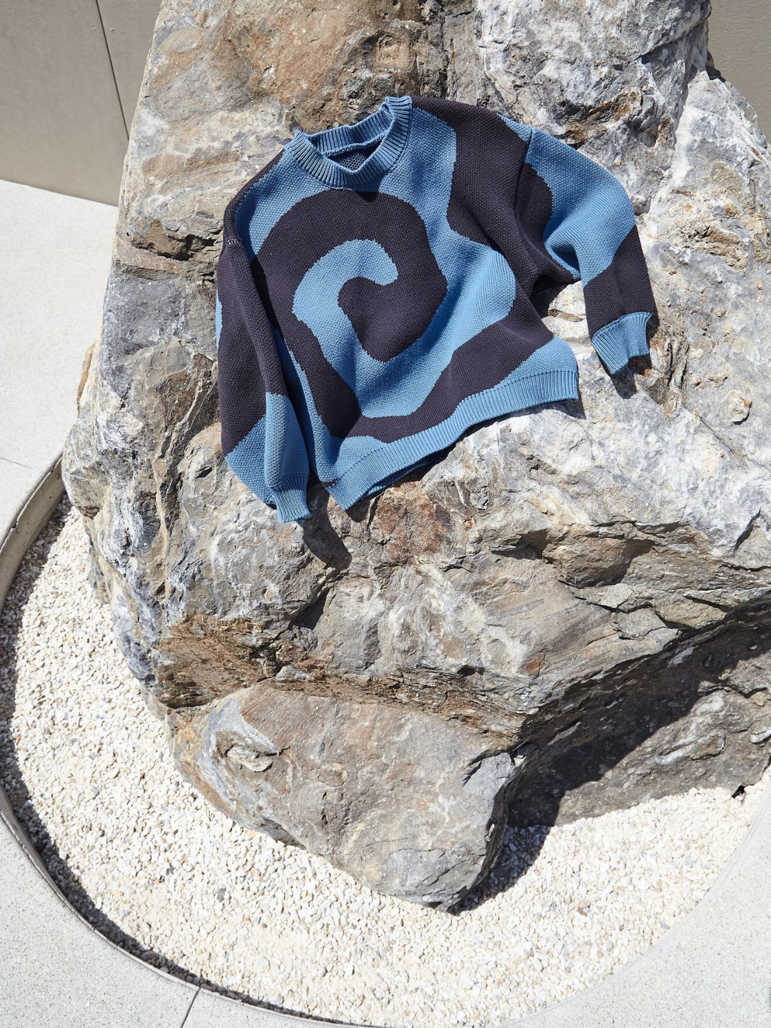 Slate | A kids crewneck sweater in medium blue, with a large single charcoal grey swirl design that covers the entire sweater, shown lying on a rock in a paved courtyard.