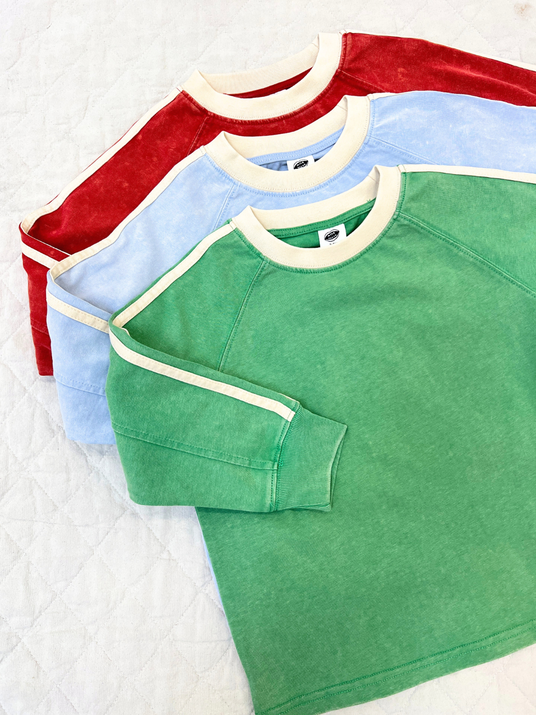Three HALFTIME LONGSLEEVE tees in green, light blue, and red with cream trim and stripe details are displayed on a quilted white surface, capturing a vintage vibe.