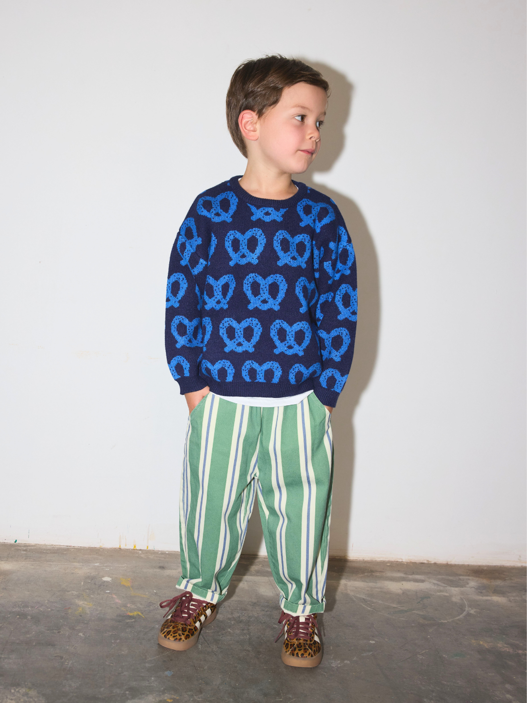 Blue | A young boy wearing a navy blue knit sweater with a blue pretzel pattern, paired with green and white vertically striped pants and brown shoes, standing against a plain white wall.