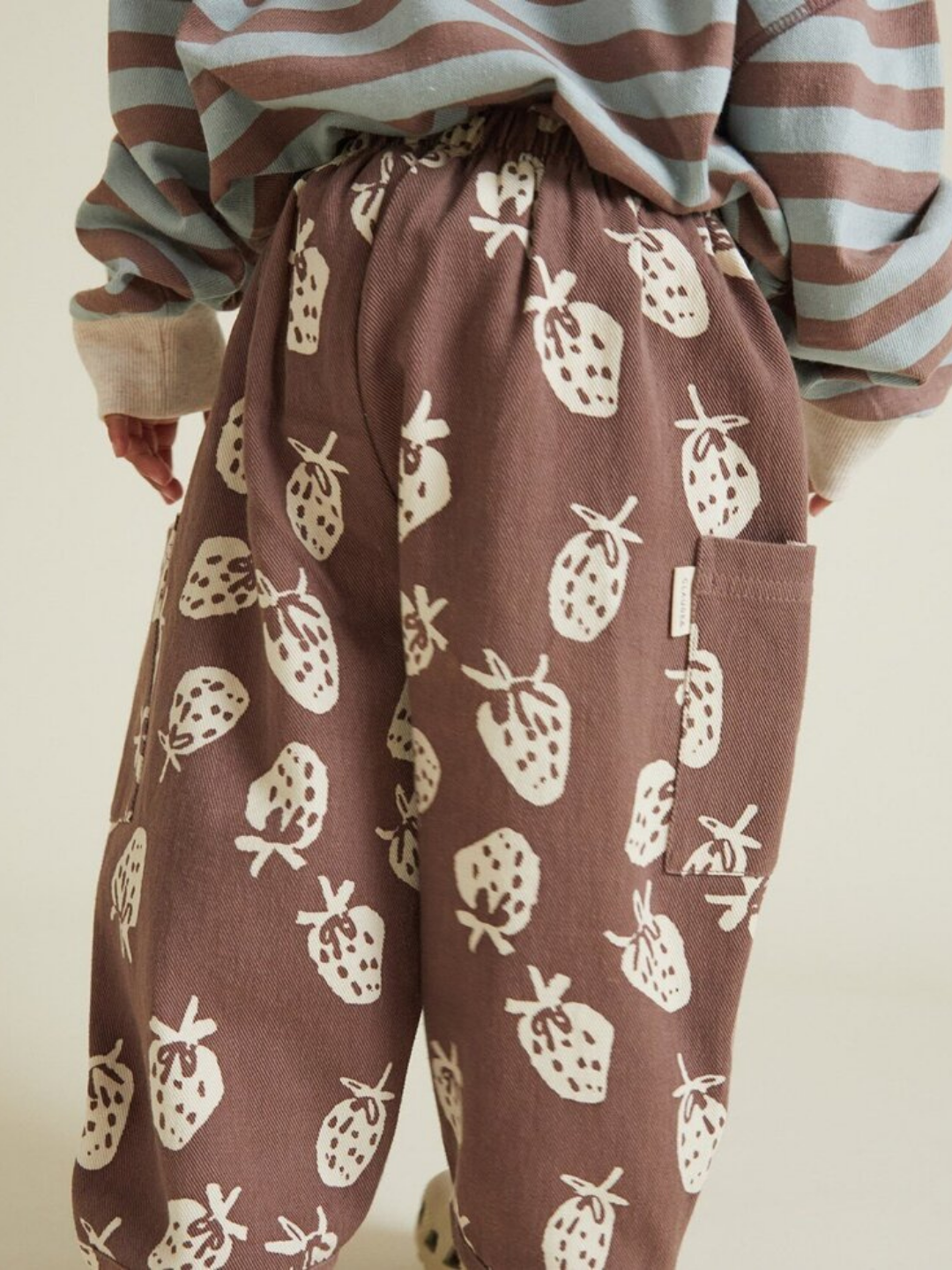 Person wearing EMERSON CHOC STRAB TROUSER, featuring a white strawberry print, paired with a blue and beige striped sweater, seen from behind.