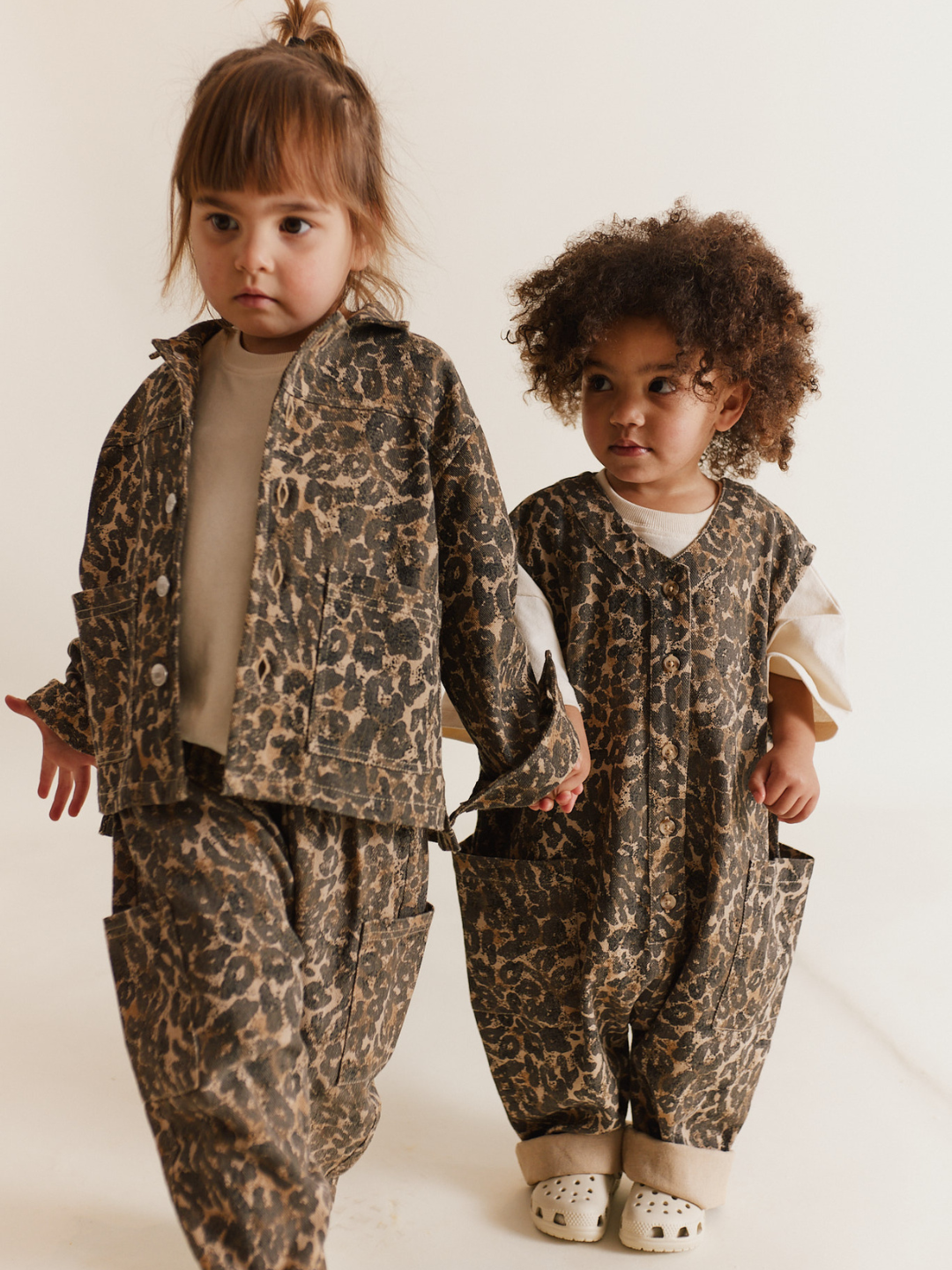 Two young children walk hand in hand on a neutral background, both wearing matching WREN LEOPARD OVERSIZED OVERALLs made from organic cotton by a unisex British brand.