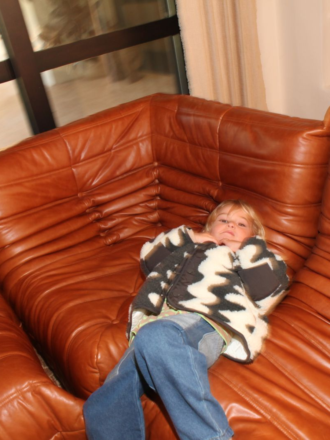 A young child wearing the WAVELENGTH SHERPA JACKET with a cozy quilted interior and jeans lies on a brown leather sectional sofa, looking up at the camera.