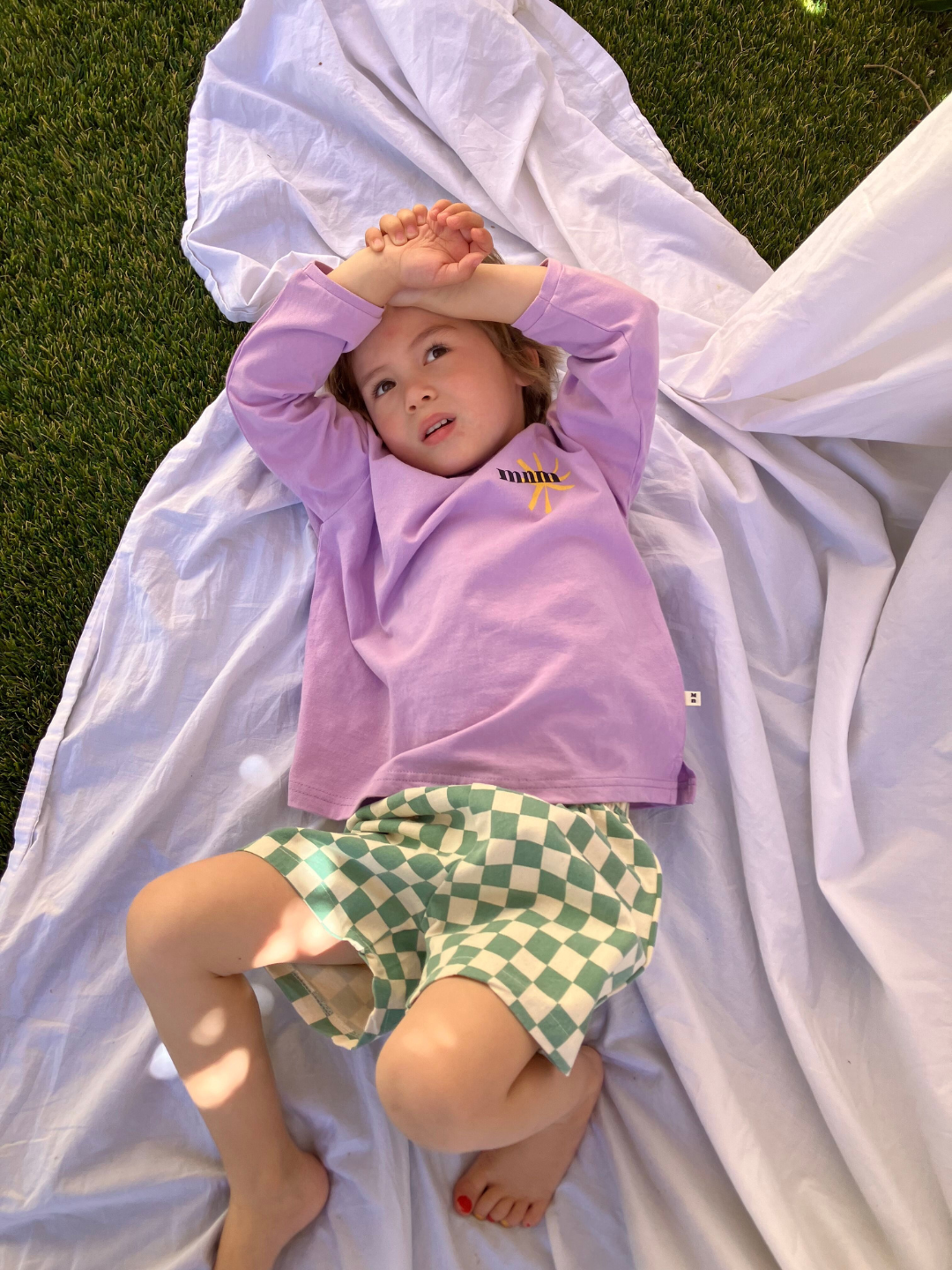 A child wearing the kids Minimal Longsleeve Tee in purple, paired with checkerboard patterned shorts in teal and ivory. He is lying on a white picnic cloth on grass.