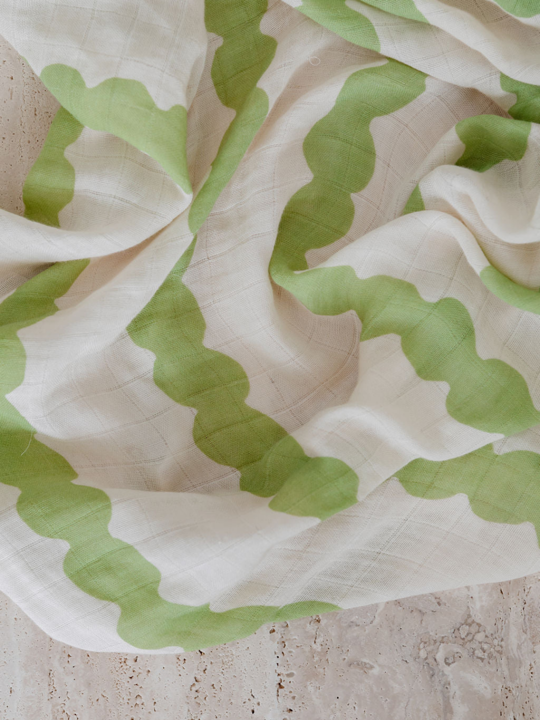 Edamame | Close-up of the ORGANIC BAMBOO SWADDLE WRAP with a pattern of green wavy stripes on a beige background, crumpled and laying on a textured stone surface. The texture and design evoke the soft comfort typical of bamboo muslin swaddles, perfect for those who appreciate quality and subtle elegance.