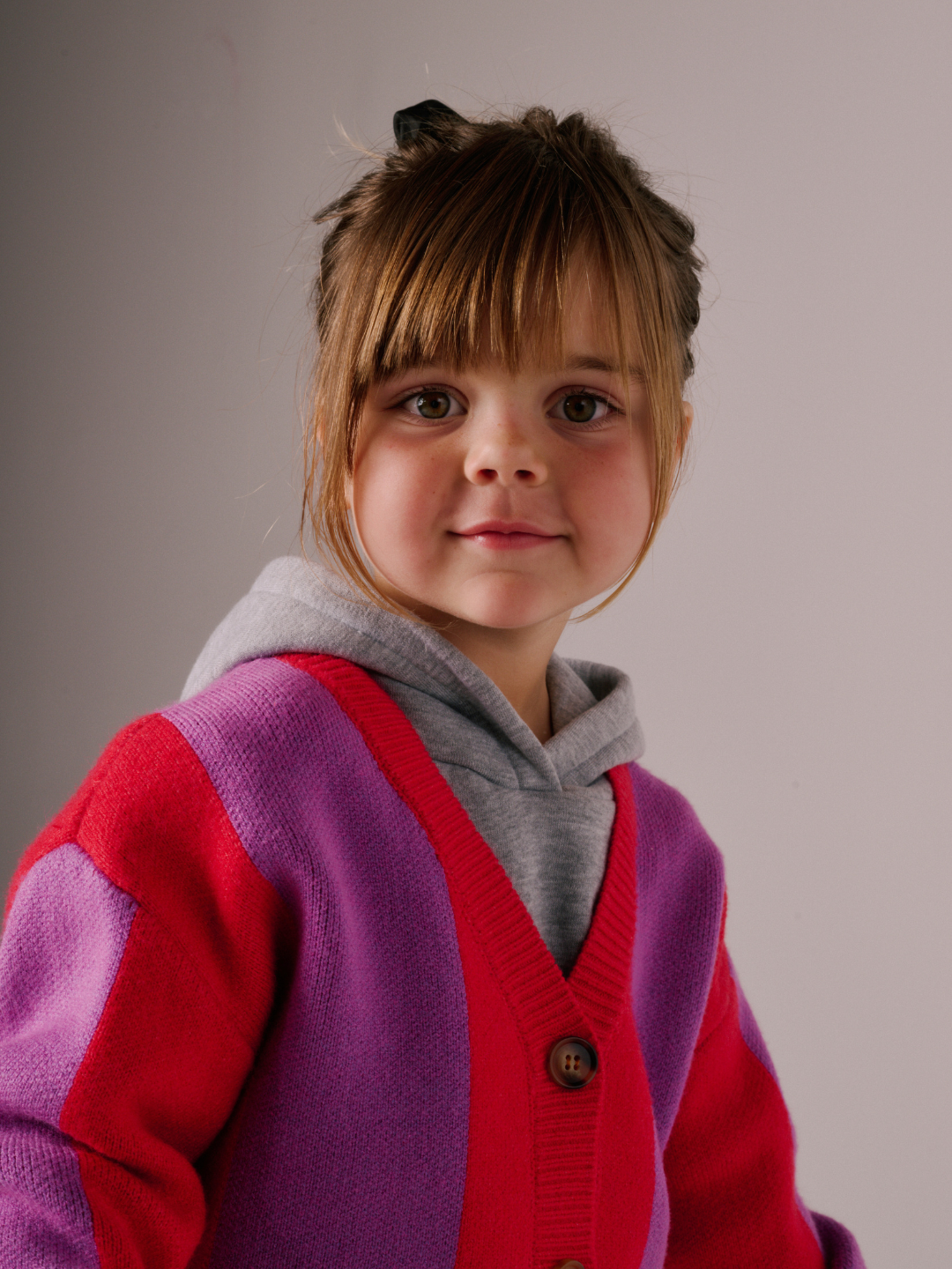 Red | A young child with brown hair wears the STRIPE CARDIGAN over a gray hoodie, standing in front of a plain background.