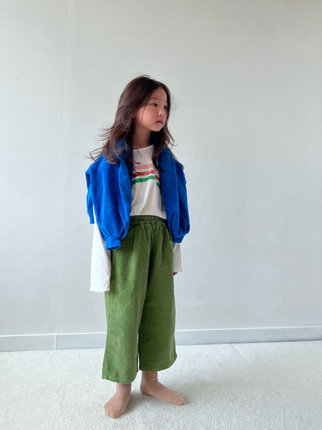 Moss | A girl wearing kids wide-leg corduroy pants in moss green, paired with a white t-shirt with three stripes in pink, red and green. A royal blue sweater is draped over her shoulders, and she is standing on grey carpet with a white wall behind her.