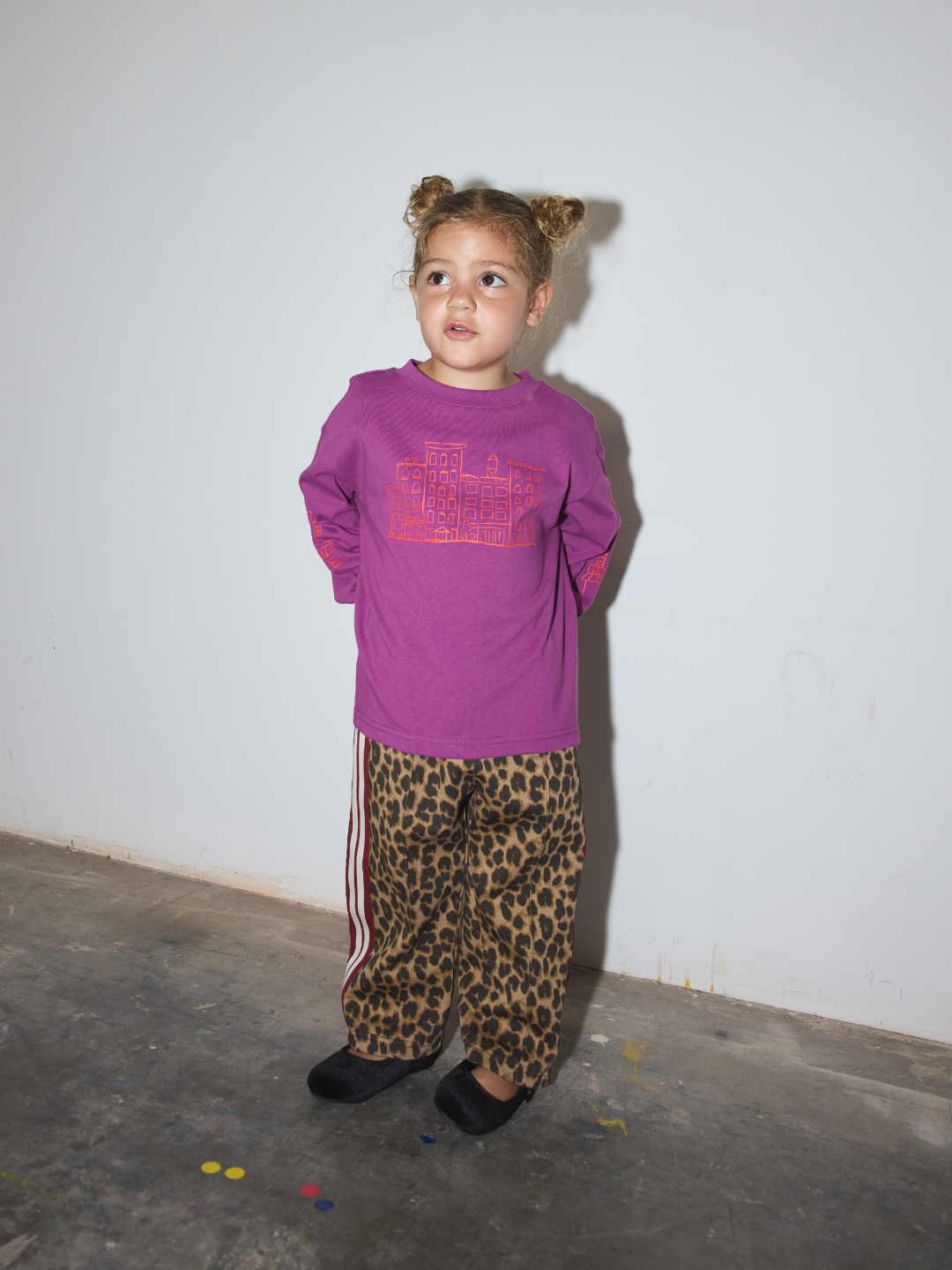 A young girl with her hair in two buns stands against a plain white wall wearing a purple long-sleeve t-shirt with a pink graphic and leopard-print pants with white side stripes.