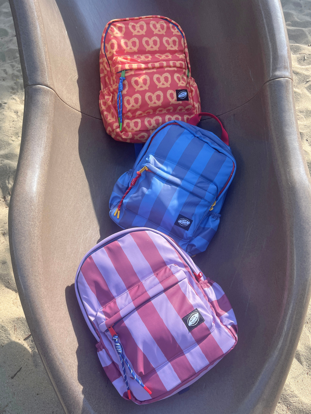 Purple Stripe | Three kids backpacks in red, blue, and purple striped patterns, arranged on a brown plastic slide outdoors on sand.