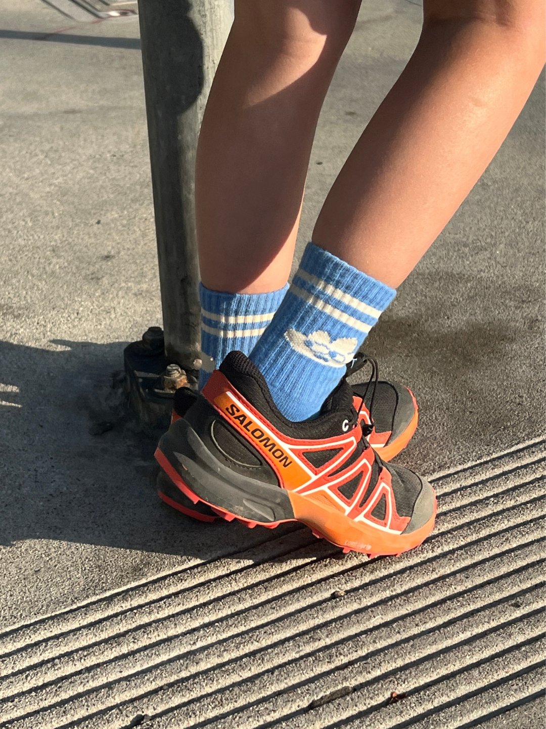 Cloud | Person wearing blue socks with white stripes and black and orange Salomon shoes on a concrete surface.