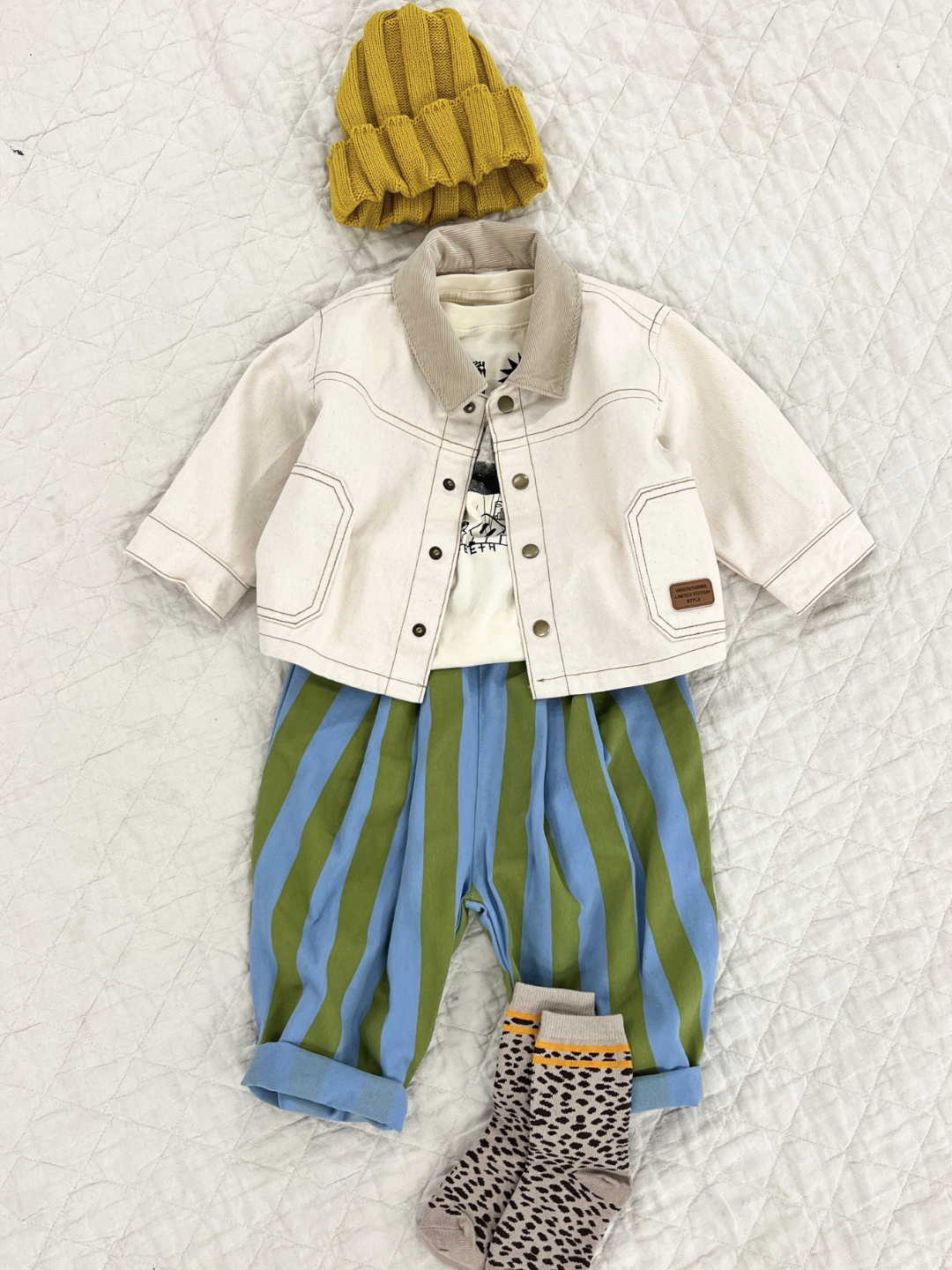Front view of kids ecru denim jacket with beige corduroy collar. Jacket is shown open over a white t-shirt, and paired with a blue and white striped pant, a yellow beanie, and cheetah print socks.