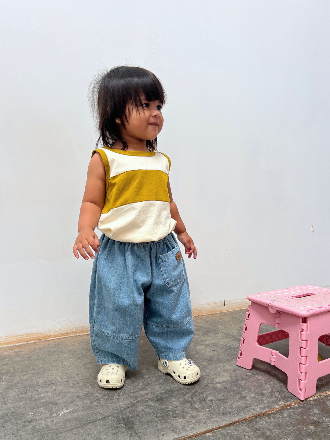 Cream/Gold | A toddler in the TRACK TANK, paired with loose blue jeans and white shoes, stands near a small pink plastic stool on a gray floor against a plain white wall.