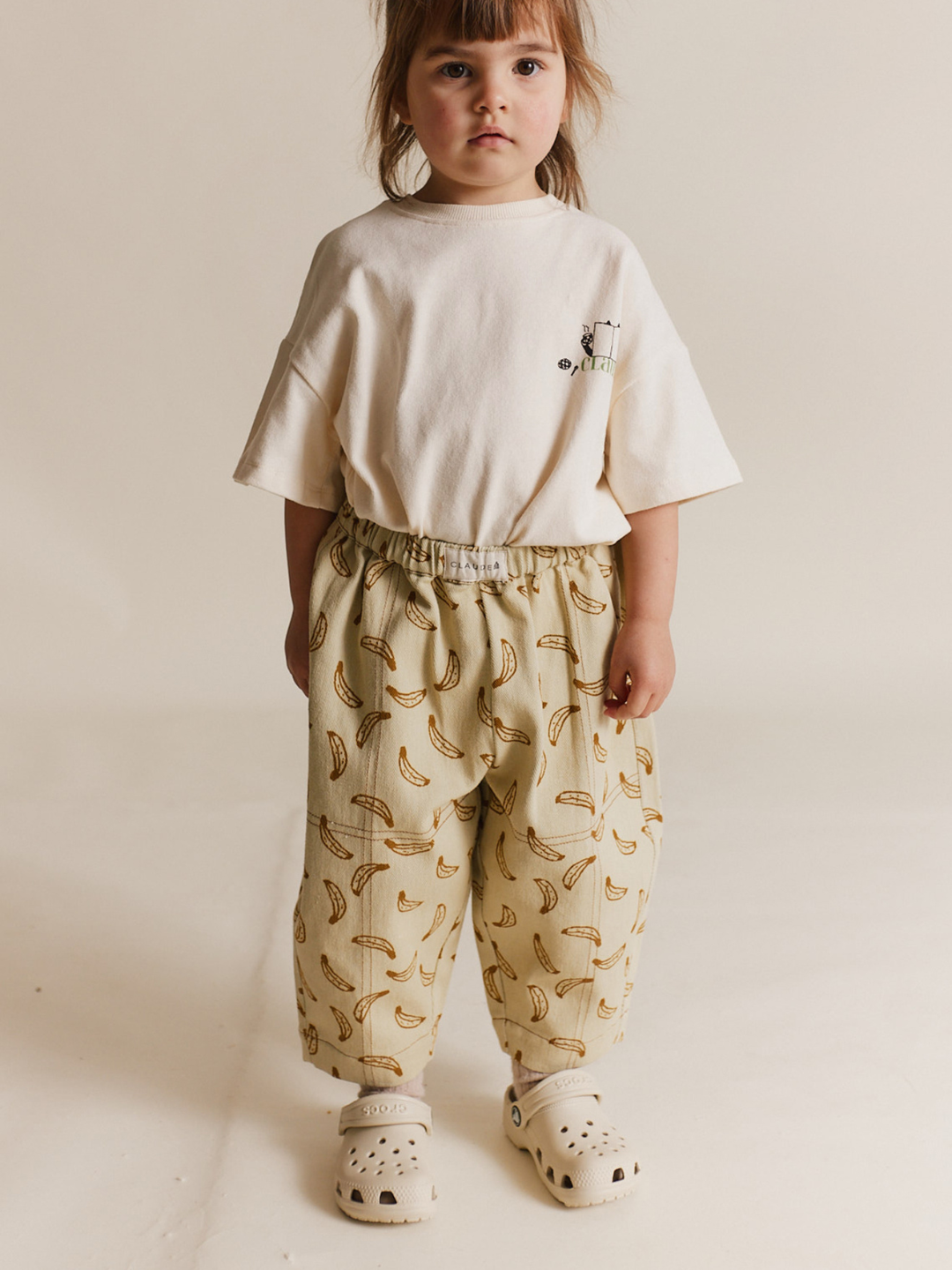 A young child stands against a plain background, wearing a cream t-shirt, white clogs, and the RIAN BANANA TROUSER—unisex pants made from banana-print organic cotton twill.