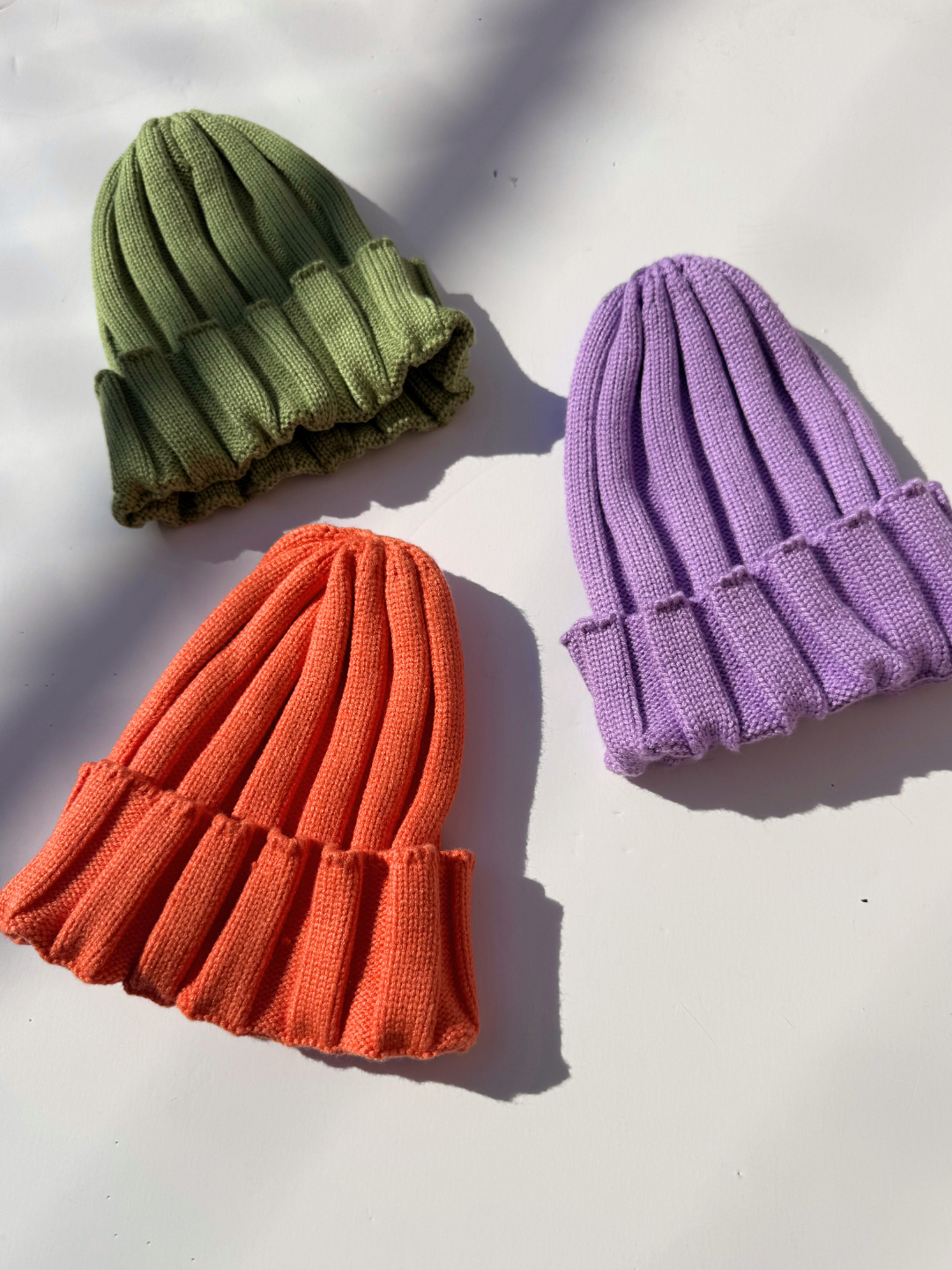 Three knit hats in green, orange, and purple on a light background