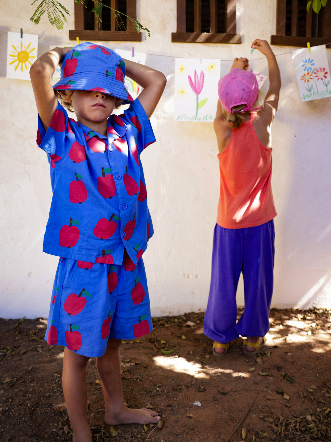 Two children hang drawings on a wall. The child in the foreground sports an APPLE SUMMER SHIRT, while the child in the background wears a pink hat, orange top, and purple pants.