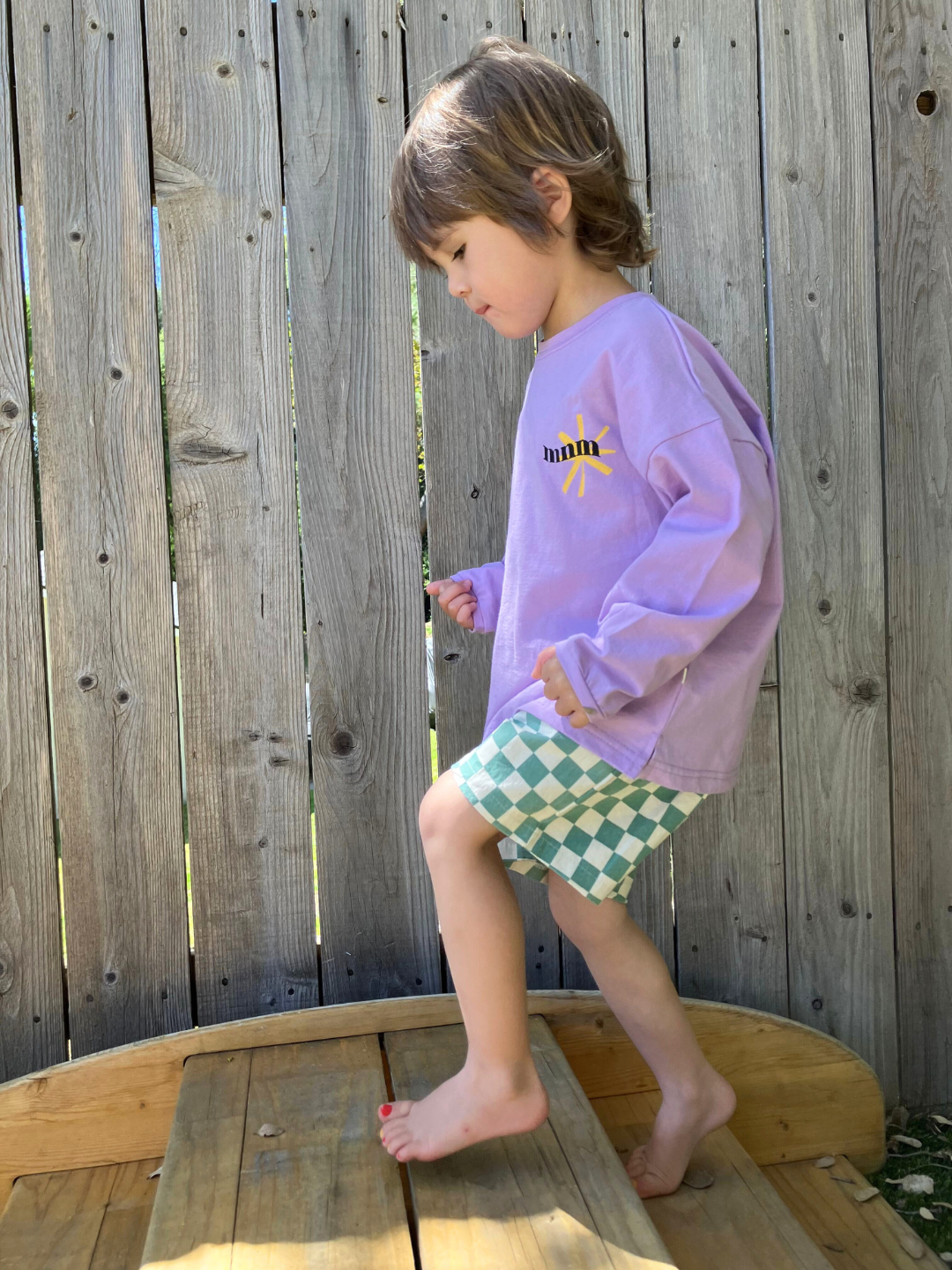 A child wearing the kids Minimal Longsleeve Tee in purple, paired with checkerboard patterned shorts in teal and ivory. He is clibing a small wooden step in front of a wooden fence.