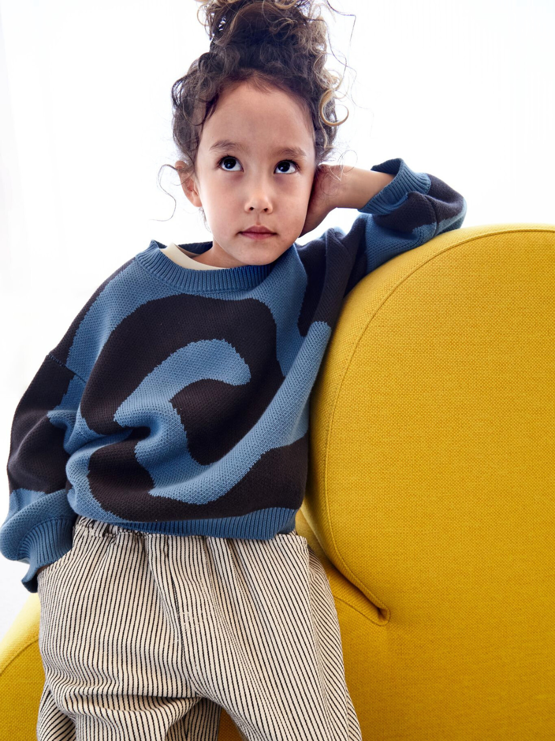 A child wearing a kids crewneck sweater in medium blue, with a large single charcoal grey swirl design that covers the entire sweater. She is seated on a yellow chair and wearing white pants with a thin black stripe.