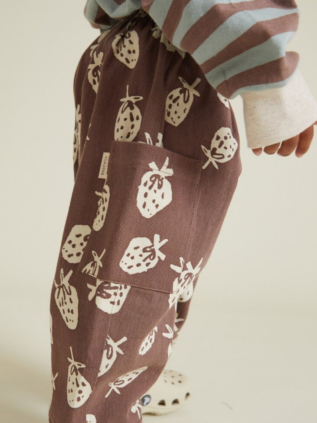 A child in EMERSON CHOC STRAB TROUSER featuring a white strawberry print and a blue-white striped top, photographed from the side.