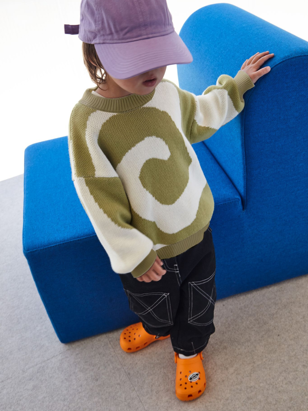 A child wearing a kids crewneck sweater in sage green, with a large single cream swirl design that covers the entire sweater. He is standing in front of a blue chair, on grey carpet. He wears black jeans with white stitching, orange shoes, and a purple cap.