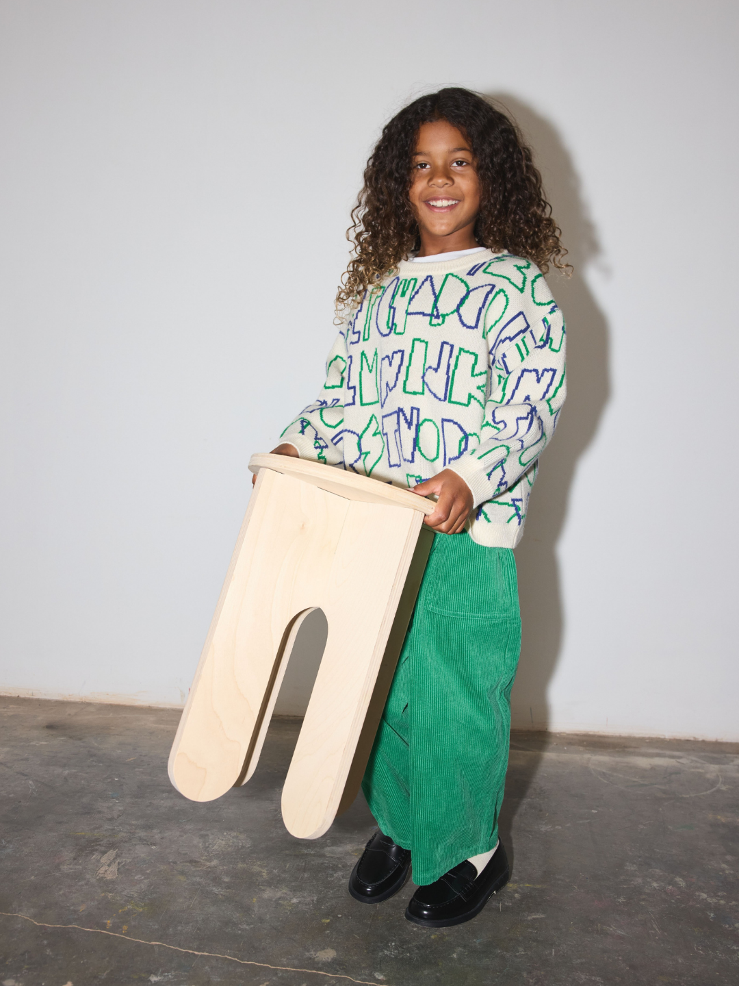 A smiling young girl with curly hair stands in a studio holding a large, light-colored wooden U-shaped block. She is wearing a white long-sleeve sweatser with green and blue abstract alphabet letters, bright green corduroy pants, and black loafers, against a plain light gray wall.