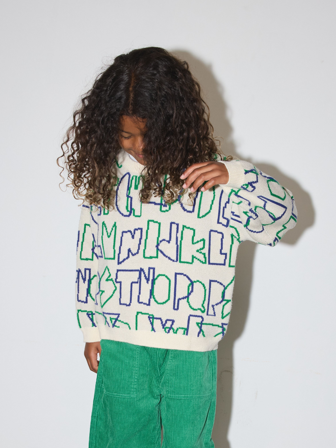 A child with long curly dark hair wearing a cream knit sweater with green and blue outlined lettering and bright green corduroy pants, standing against a plain light gray wall.