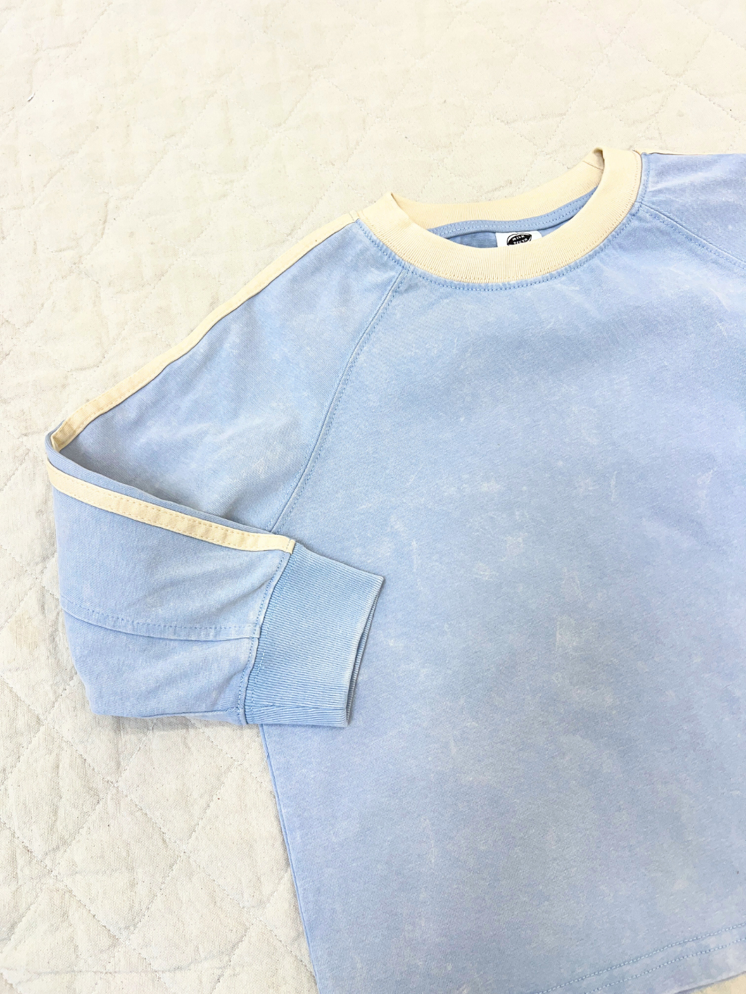 Blue | The HALFTIME LONGSLEEVE is a light blue, oversized tee with beige trim on the collar, cuffs, and sleeve top, styled laid flat on a quilted cream surface for a cozy vintage vibe.
