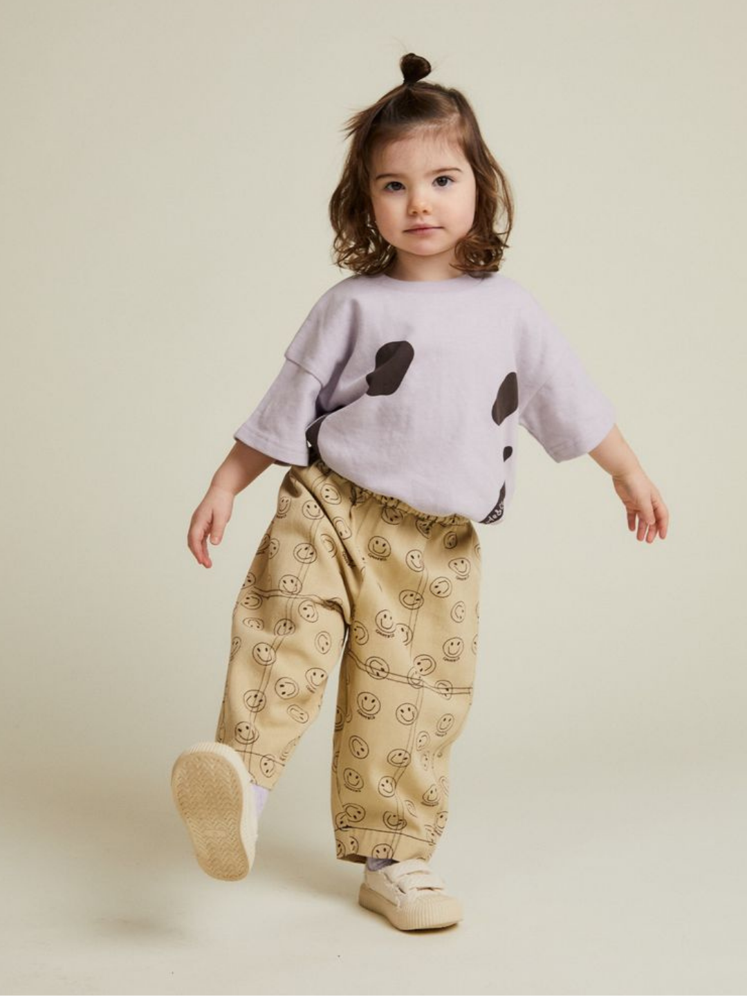A young child indoors wears REX JOY BUTTER TROUSER—beige pants with yellow smiley faces—paired with a light purple shirt with black spots, looking at the camera.