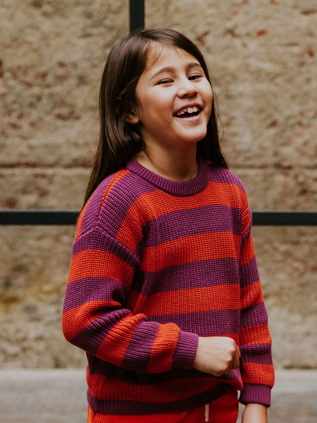 Lollipop | A young girl with long brown hair, wearing the STRIPES SWEATER in red and purple stripes, smiles outdoors against a textured stone wall.