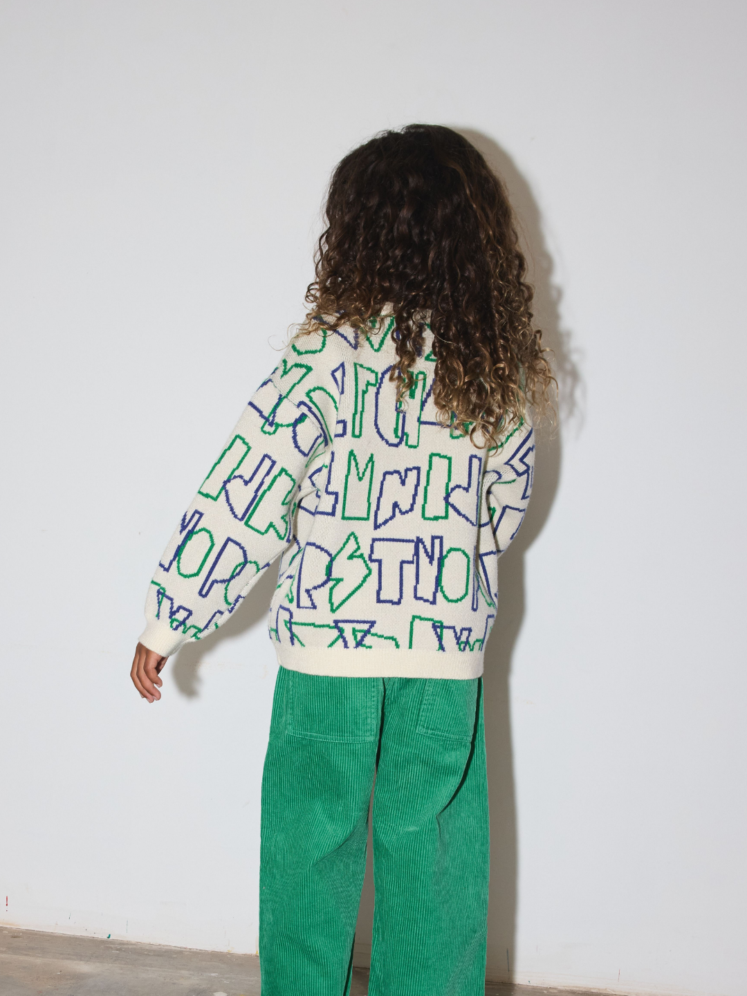 A child shown from the back wearing a cream-colored long-sleeve sweater with abstract green and blue line-art text patterns, paired with green corduroy pants, standing against a plain white wall.