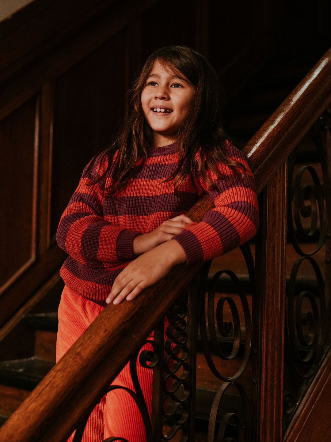 Lollipop | A young girl with long hair stands on a staircase, leaning on the wooden handrail and smiling. She is wearing the STRIPES SWEATER in red along with matching red pants.