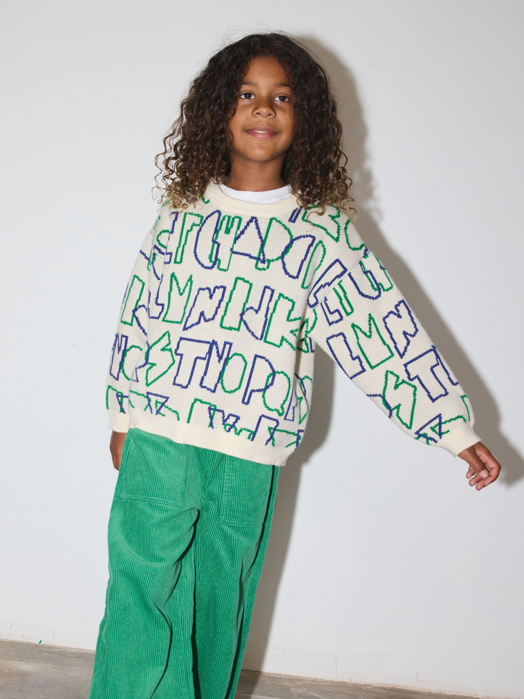 A child with curly hair wearing a cream-colored crewneck sweatshirt with a green and blue graphic text pattern, paired with green corduroy pants, standing against a plain white wall.