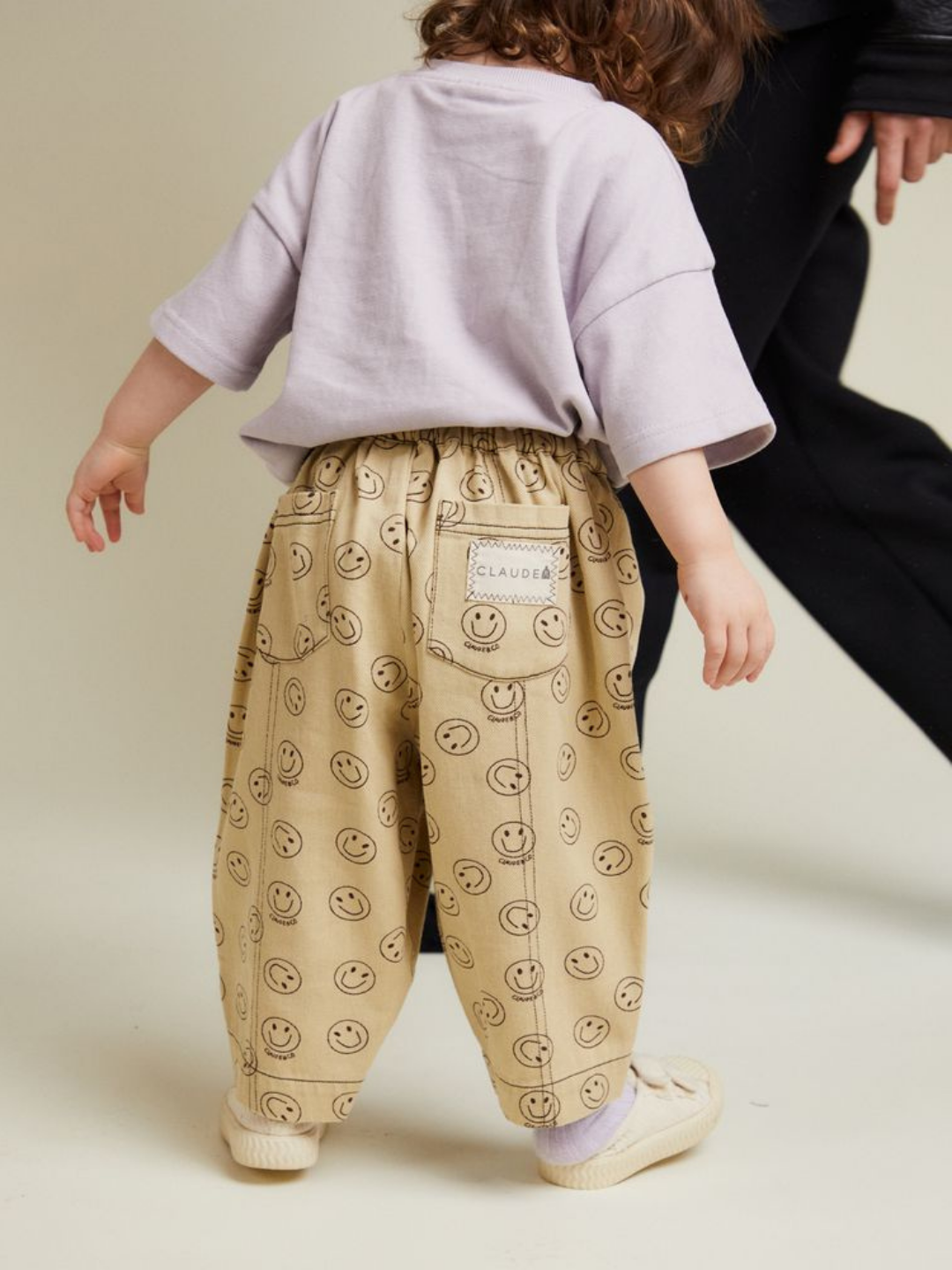 A young child in a loose light purple T-shirt and REX JOY BUTTER TROUSER walks beside an adult dressed in black.