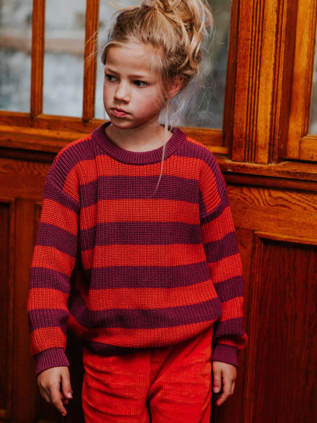 Lollipop | A young child with blonde hair, wearing the STRIPES SWEATER featuring red and dark purple stripes, stands in front of a wooden door, looking to the side.