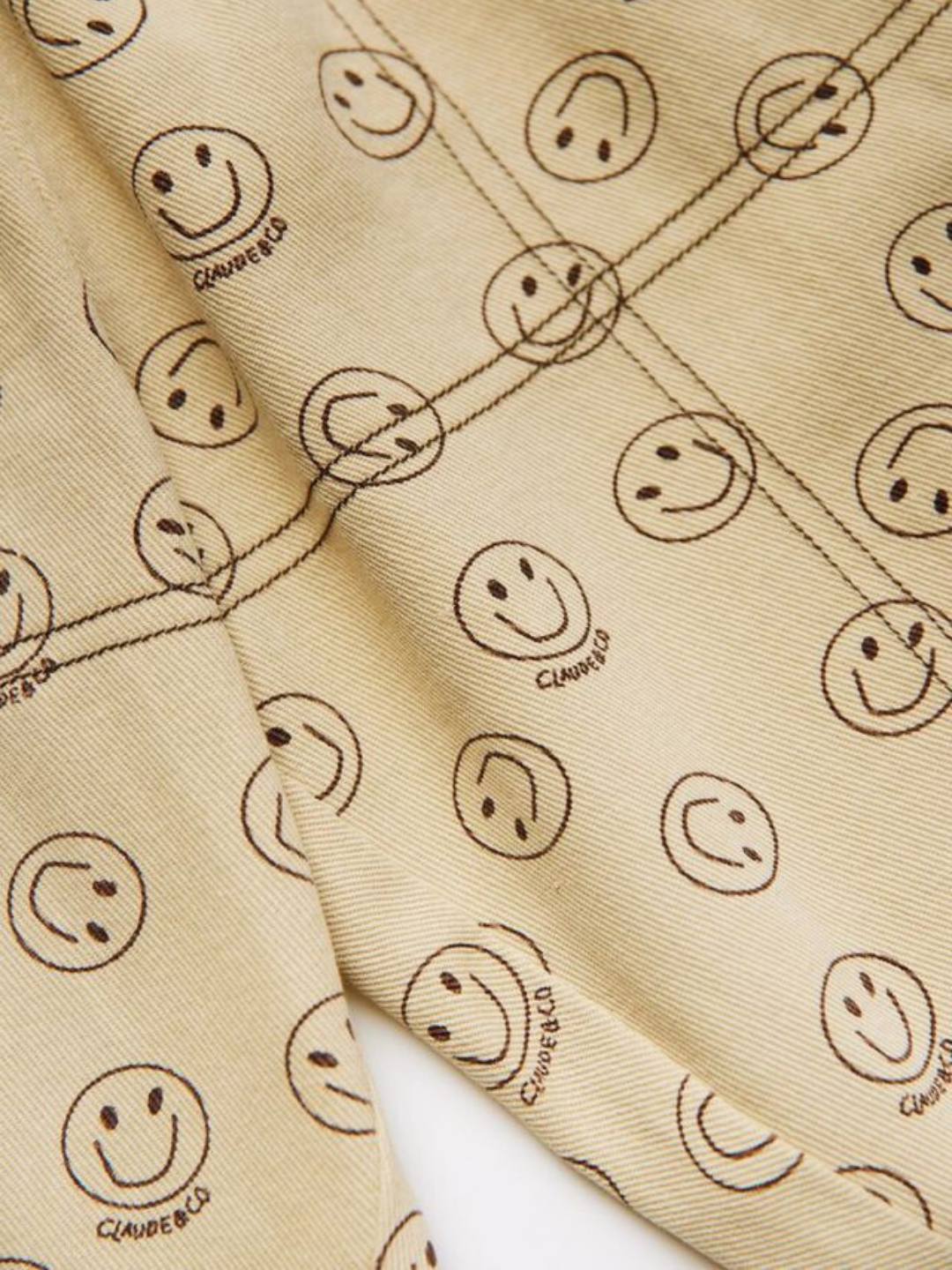 The REX JOY BUTTER TROUSER features beige fabric decorated with a pattern of brown smiley faces and "CLAUDE & CO" printed beneath some of them.