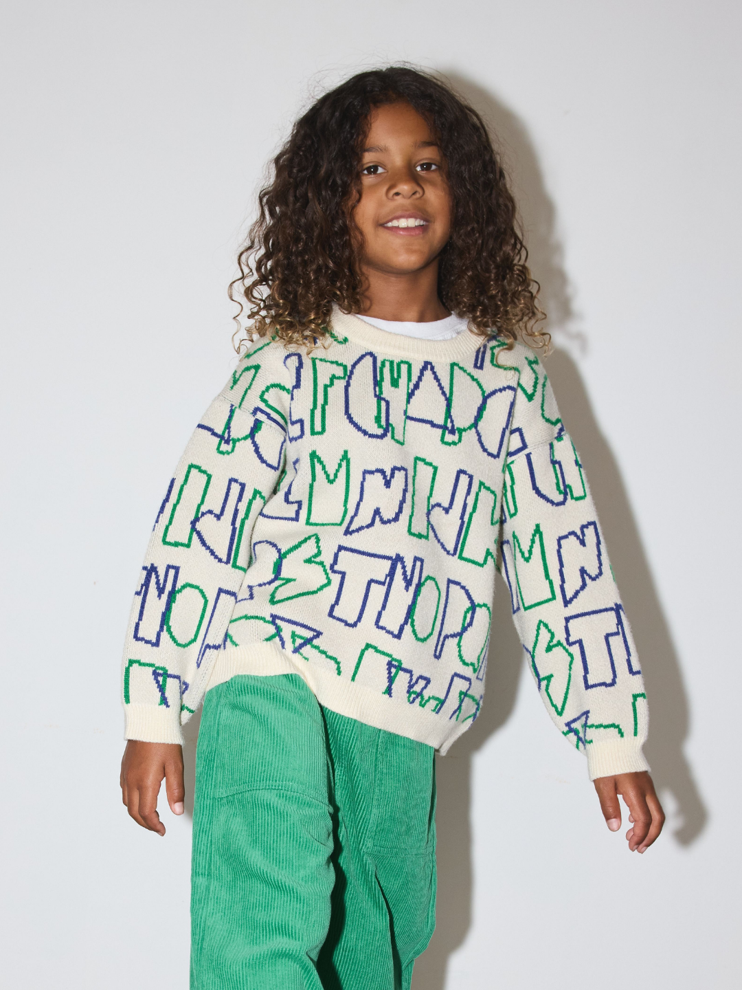 A young girl with curly brown hair wearing a cream-colored knit sweater with a green and blue graffiti-style letter pattern, paired with green corduroy pants, standing against a light gray studio background.