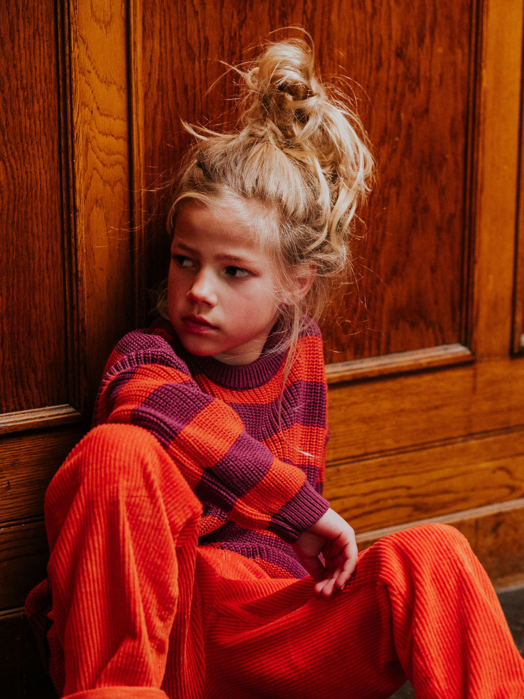 Lollipop | A child with blonde hair in a messy bun, wearing the STRIPES SWEATER featuring red and purple stripes, paired with orange pants, and sitting against a wooden wall looking to the side.