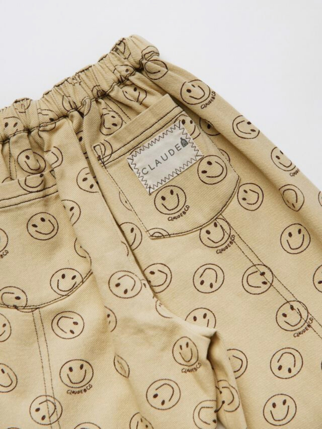 The REX JOY BUTTER TROUSER are beige pants with an elastic waistband, featuring a repeating black smiley face pattern and a stitched "CLAUDE" label.