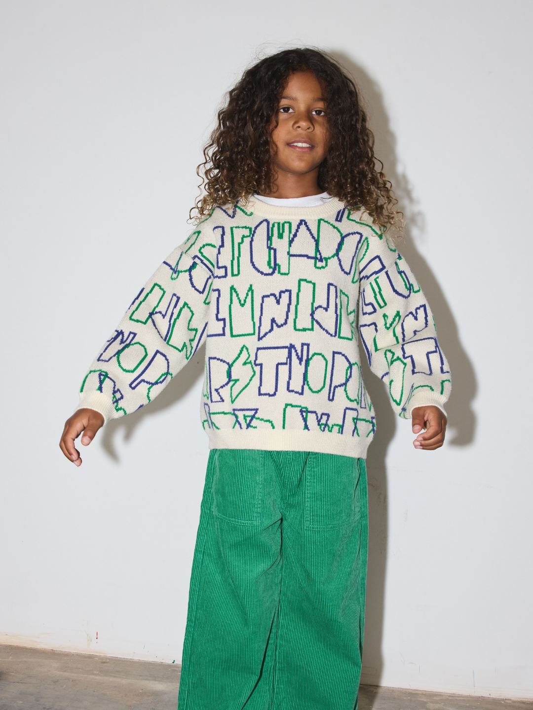A child wearing a cream-colored knit sweater with a colorful graffiti-style text pattern in green and blue, paired with green wide-leg pants, standing against a plain white wall.