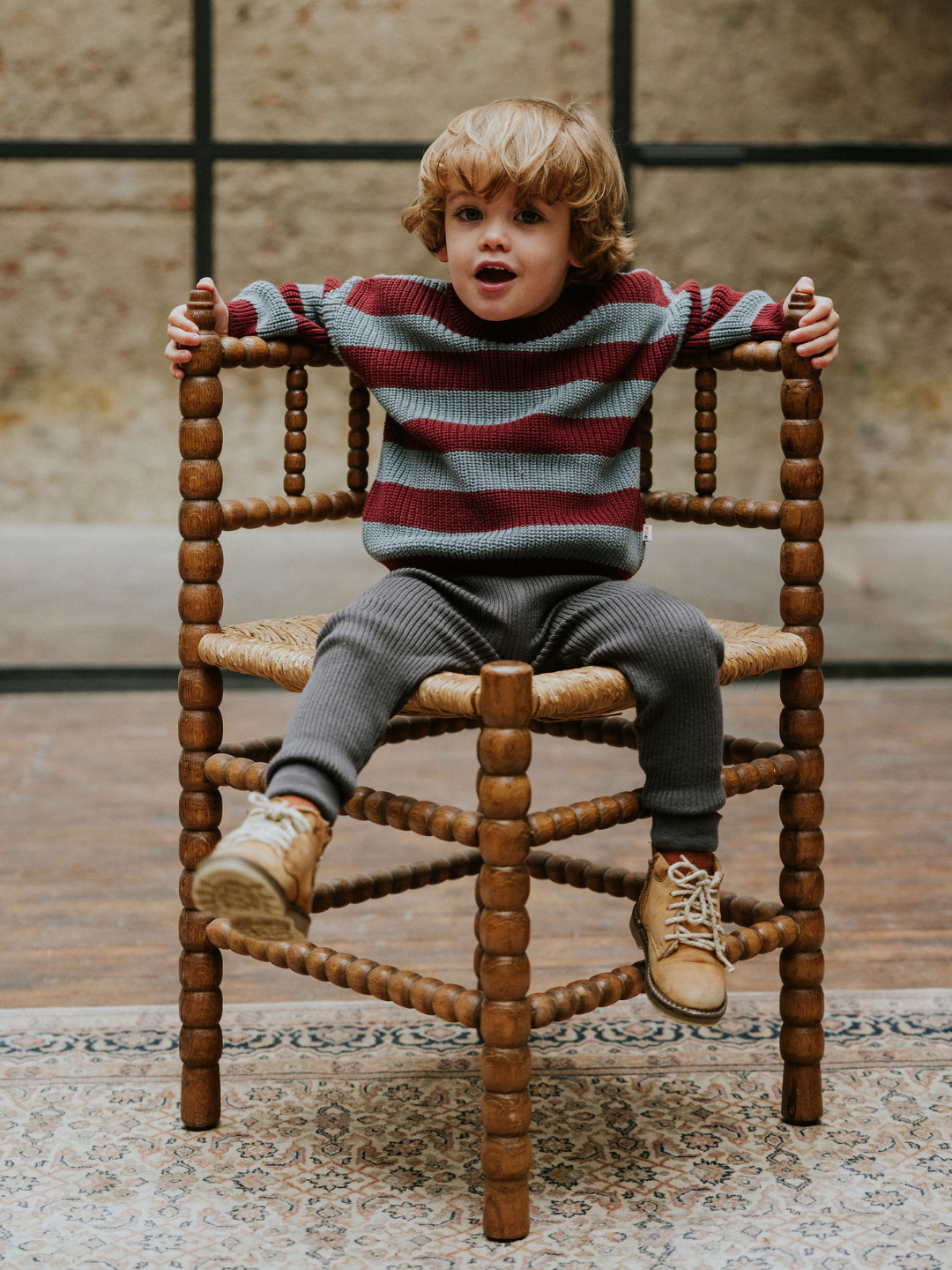 Seahorse | A young child with curly hair sits on a wooden chair with both hands on the armrests. They are wearing a red and gray STRIPES SWEATER, gray pants, and beige sneakers.