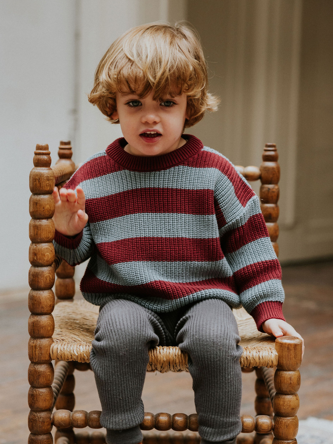 Seahorse | A young child with curly hair sits on a wooden chair, wearing the STRIPES SWEATER in burgundy and gray, and gray leggings, with one hand raised.