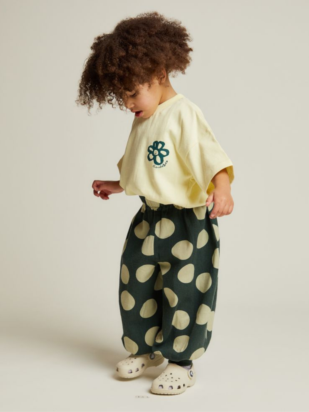 A young child with curly hair stands against a plain background wearing a yellow t-shirt with a flower graphic, white clogs, and the EMERSON KALE POLKA TROUSER—loose dark pants with large beige spots.